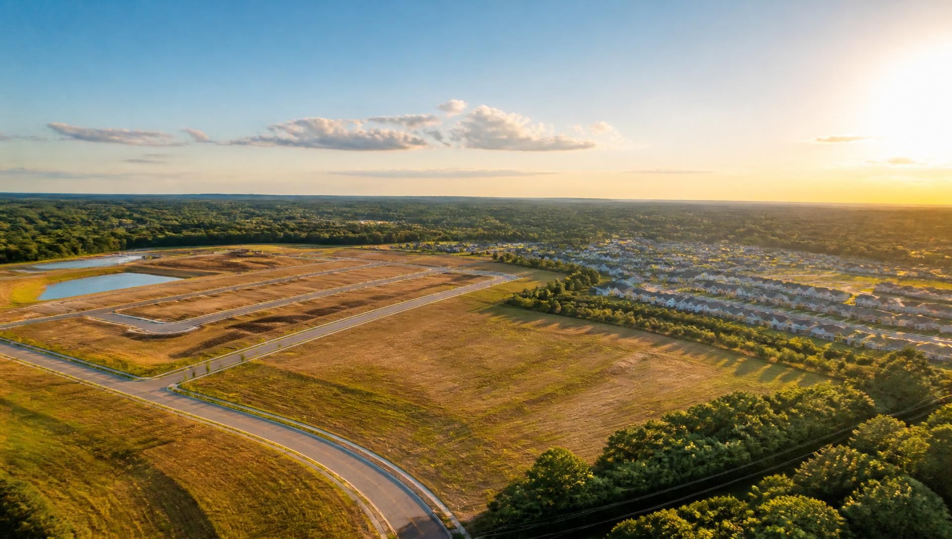 Aerial view of graded residential land parcels beside a new neighborhood at golden hour